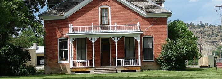 Historic red brick house with a white Victorian-style porch, surrounded by trees and a grassy lawn.