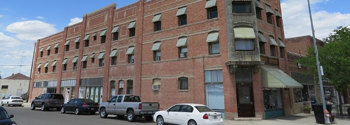 Historic multi-story red brick building with awnings, storefront windows, and parked cars along the street.