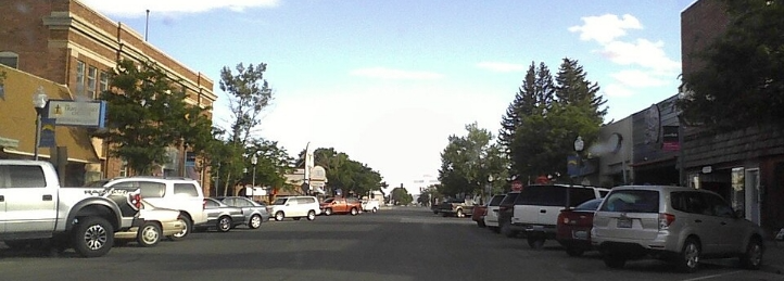 Downtown street lined with parked cars, brick buildings, storefronts, and mature trees under a partly cloudy sky.