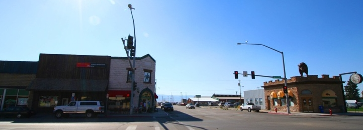 Downtown street intersection with storefronts, traffic lights, parked vehicles, and a clear blue sky.