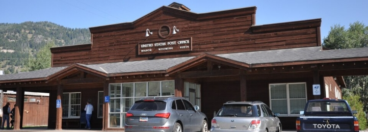 United States Post Office with a rustic wood facade, parked cars, and mountains in the background.