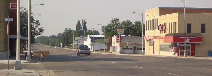 Small-town commercial street with storefronts, streetlights, and a pickup truck driving along a wide road.